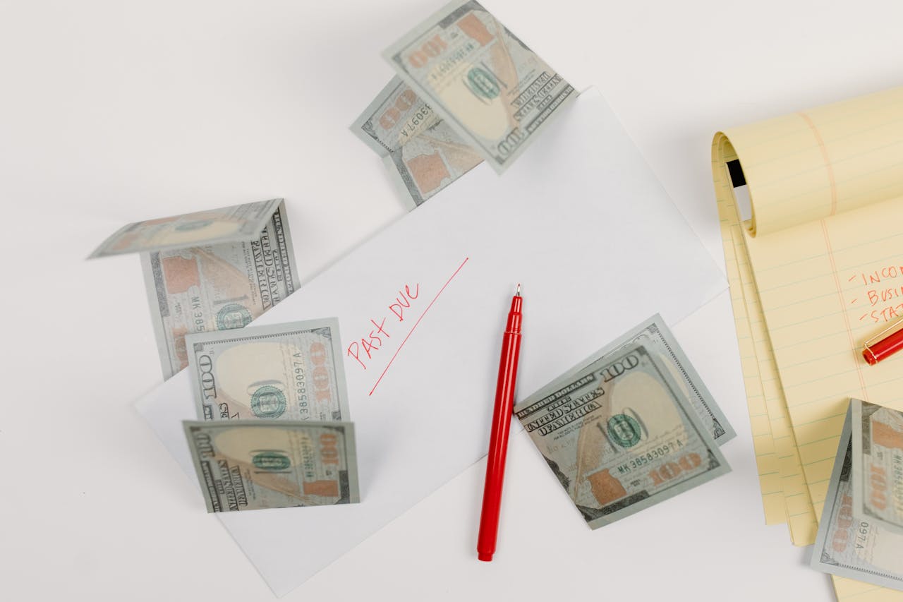 A top-down view of scattered US dollar bills with a 'past due' envelope, red pen, and notepad.